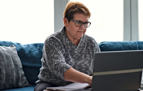 Older woman using a laptop at home while reviewing paperwork and taking notes, representing budgeting or personal finance management.