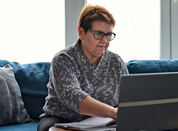 Older woman using a laptop at home while reviewing paperwork and taking notes, representing budgeting or personal finance management.