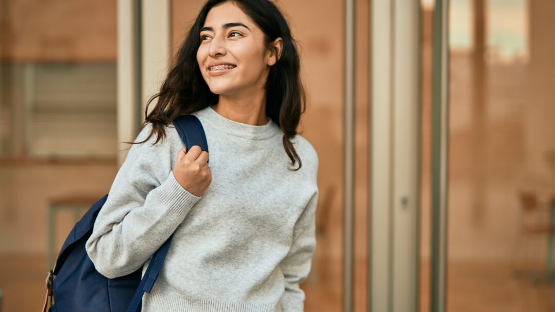 Smiling female college student wearing a backpack outside an academic building, representing higher education and campus life.