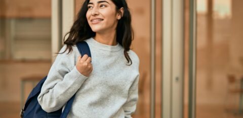 Smiling female college student wearing a backpack outside an academic building, representing higher education and campus life.