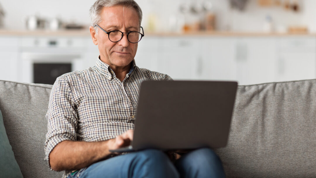Older man using a laptop while sitting on a couch at home.