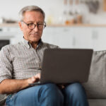 Older man using a laptop while sitting on a couch at home.