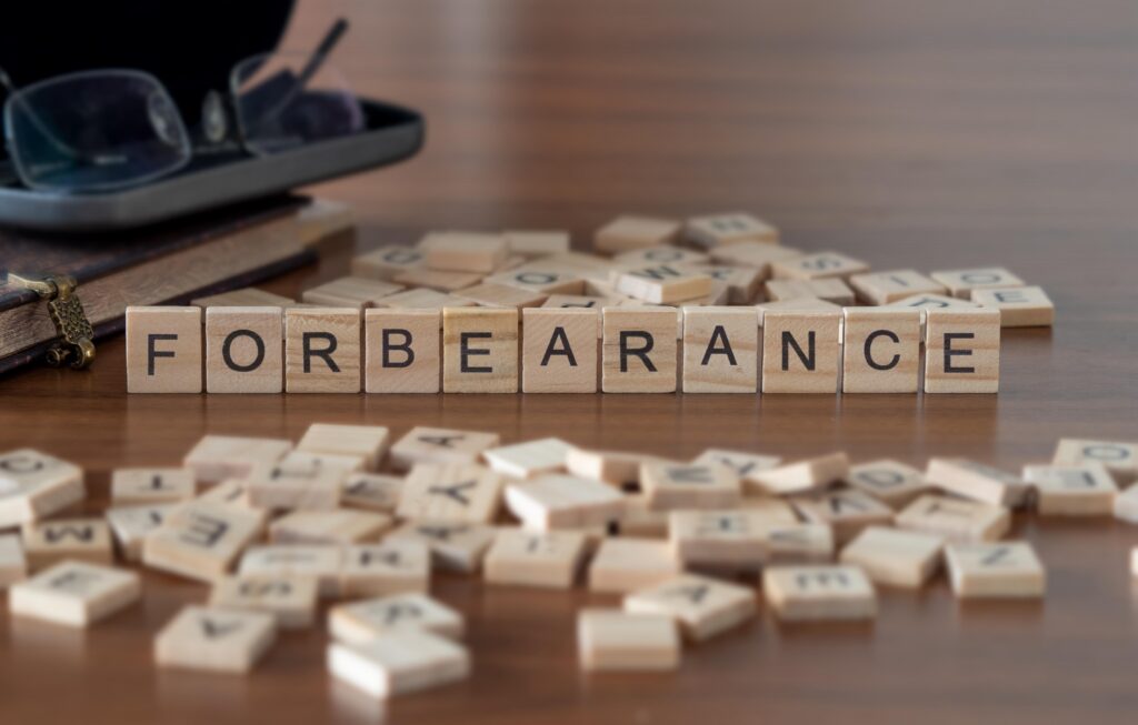 Wooden letter blocks spelling "FORBEARANCE" on a table surrounded by scattered letter tiles, books, and eyeglasses.