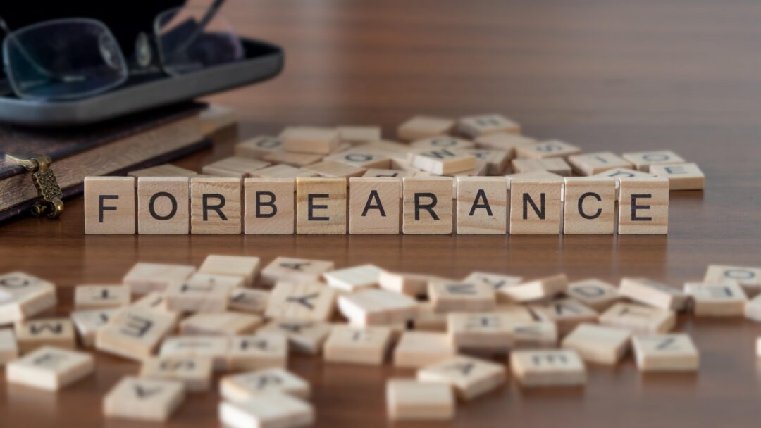 Wooden letter blocks spelling "FORBEARANCE" on a table surrounded by scattered letter tiles, books, and eyeglasses.