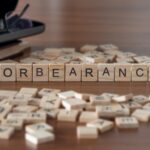 Wooden letter blocks spelling "FORBEARANCE" on a table surrounded by scattered letter tiles, books, and eyeglasses.