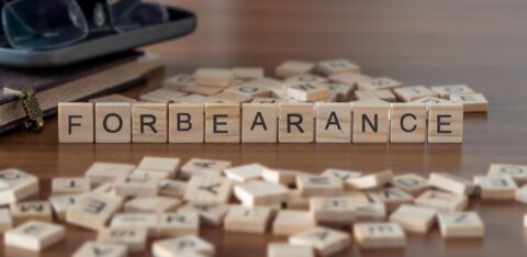 Wooden letter blocks spelling "FORBEARANCE" on a table surrounded by scattered letter tiles, books, and eyeglasses.
