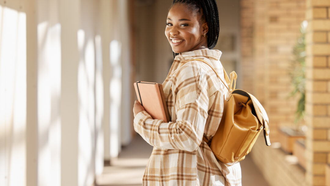 Smiling female college student carrying a backpack and holding notebooks while walking through a campus hallway.