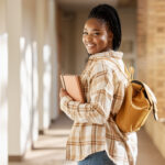 Smiling female college student carrying a backpack and holding notebooks while walking through a campus hallway.