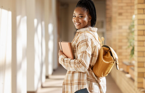 Smiling female college student carrying a backpack and holding notebooks while walking through a campus hallway.