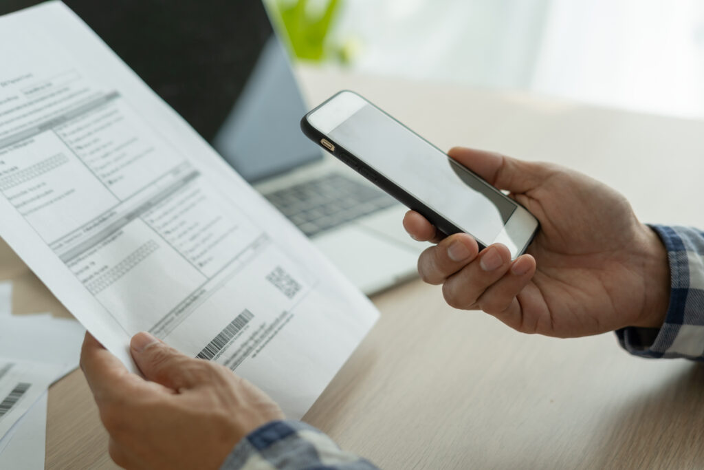 Person holding a smartphone while reviewing a bill, with a laptop in the background, representing online bill payment or account management.