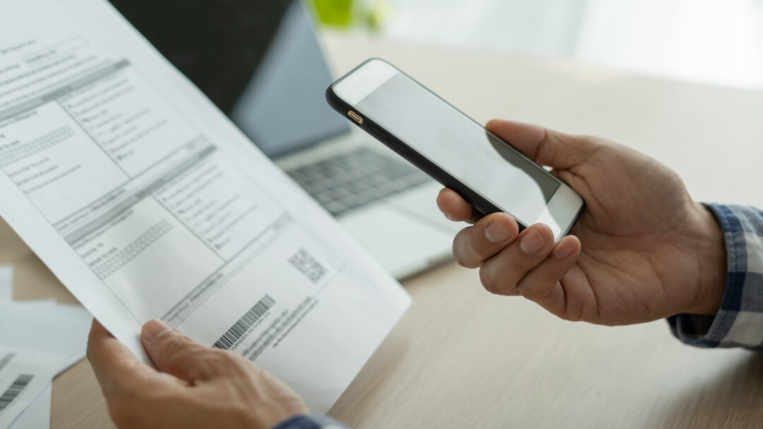 Person holding a smartphone while reviewing a bill, with a laptop in the background, representing online bill payment or account management.