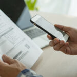 Person holding a smartphone while reviewing a bill, with a laptop in the background, representing online bill payment or account management.