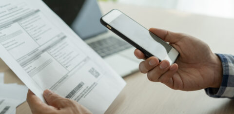 Person holding a smartphone while reviewing a bill, with a laptop in the background, representing online bill payment or account management.