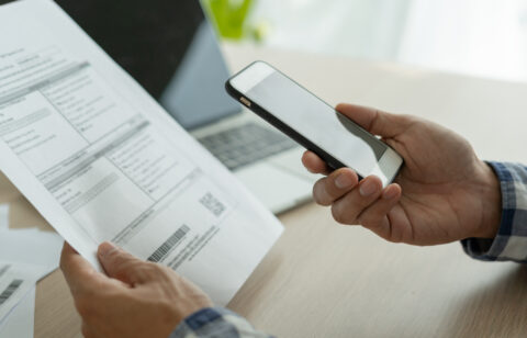 Person holding a smartphone while reviewing a bill, with a laptop in the background, representing online bill payment or account management.
