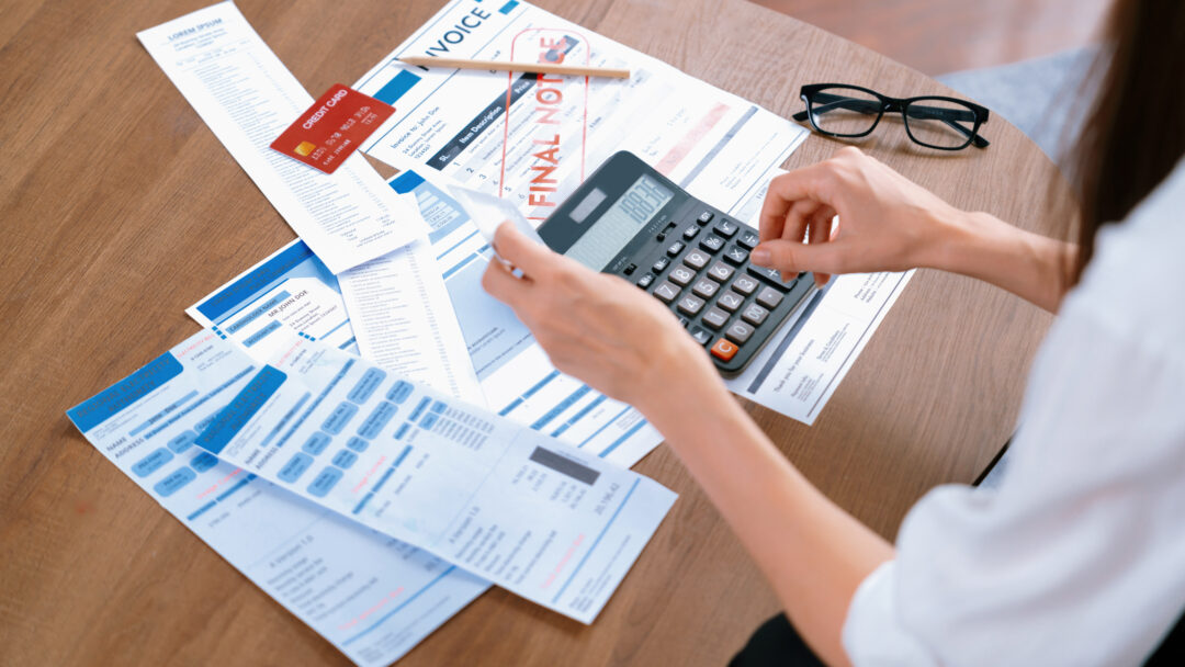 Person reviewing bills and receipts while using a calculator at a table with a credit card, invoice, and financial documents.
