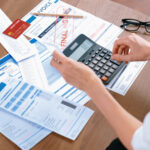 Person reviewing bills and receipts while using a calculator at a table with a credit card, invoice, and financial documents.