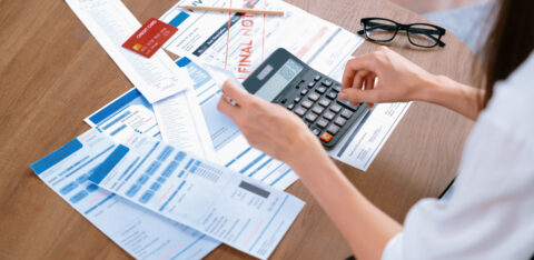 Person reviewing bills and receipts while using a calculator at a table with a credit card, invoice, and financial documents.