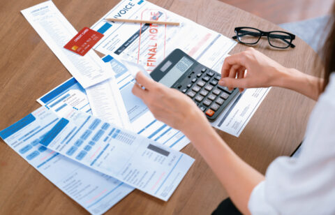Person reviewing bills and receipts while using a calculator at a table with a credit card, invoice, and financial documents.