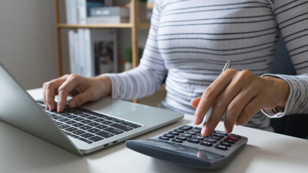 Person using a calculator while working on a laptop, holding a pen and entering numbers at a desk.