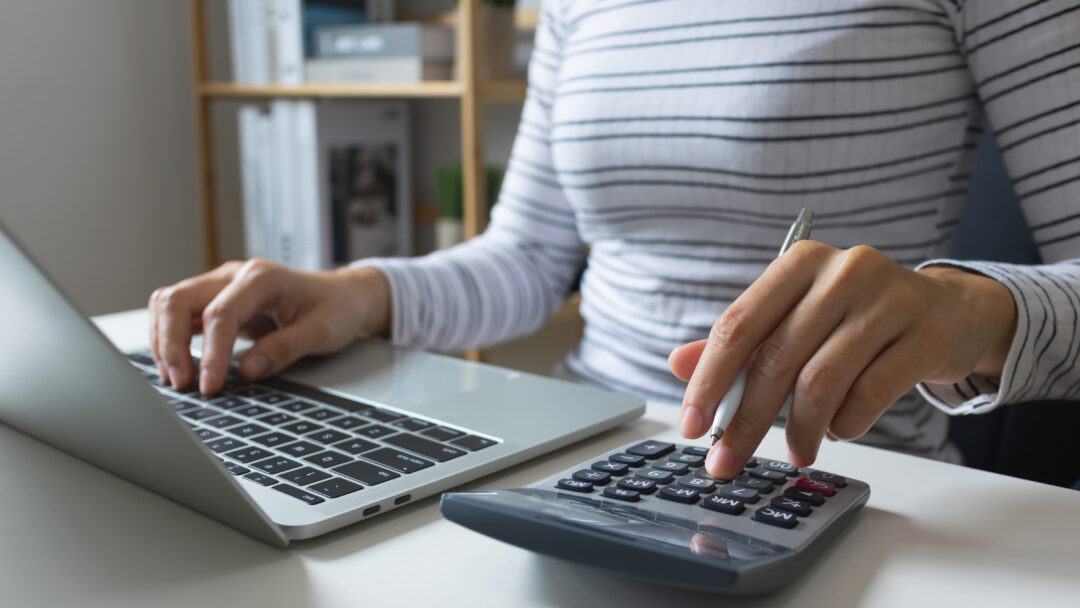 Person using a calculator while working on a laptop, holding a pen and entering numbers at a desk.