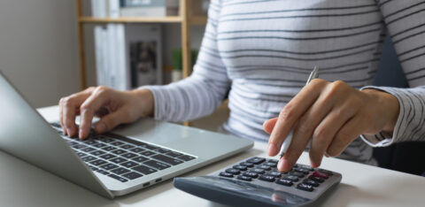 Person using a calculator while working on a laptop, holding a pen and entering numbers at a desk.