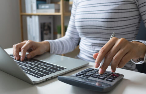 Person using a calculator while working on a laptop, holding a pen and entering numbers at a desk.
