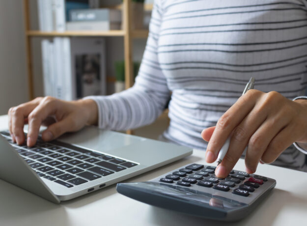 Person using a calculator while working on a laptop, holding a pen and entering numbers at a desk.