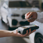 Person using a credit card to make a payment with a card reader at a car dealership, representing vehicle purchases or financing.