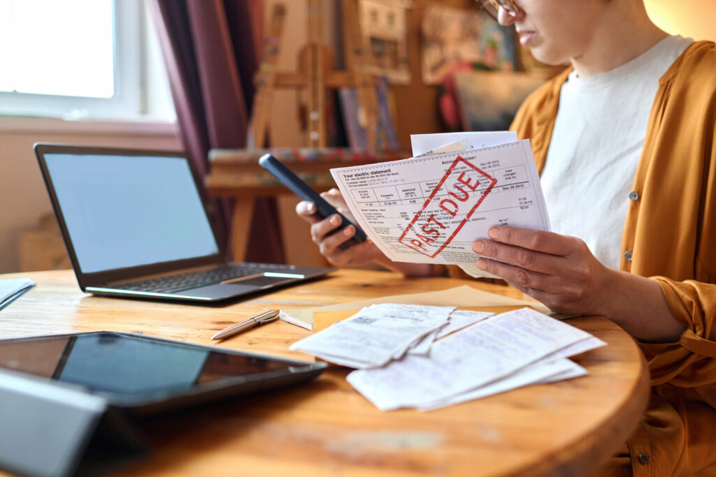 Woman reviewing an overdue bill while holding a smartphone at a table with a laptop and receipts.