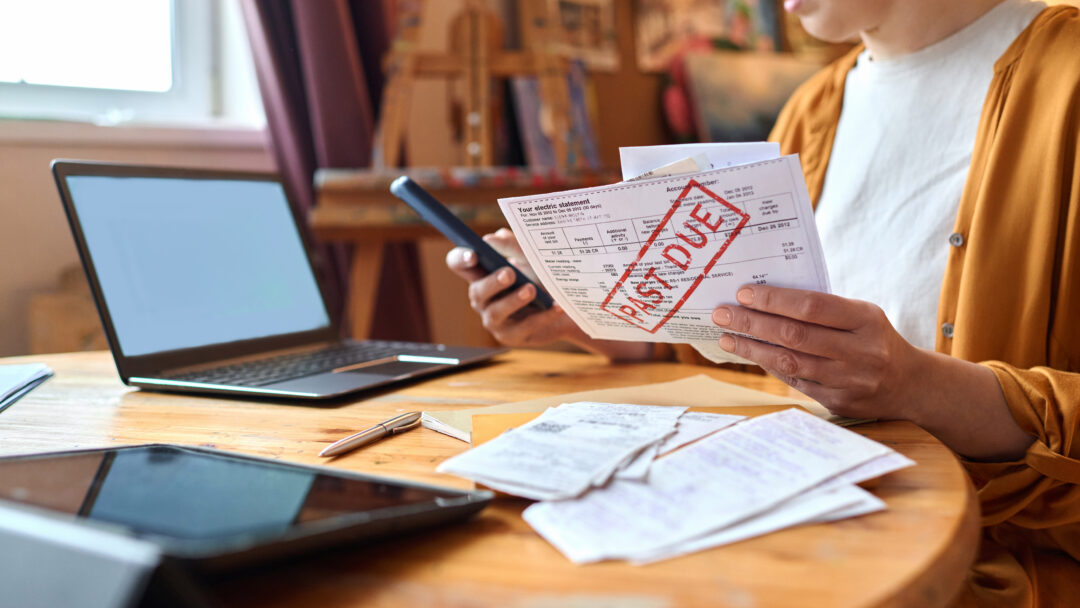 Woman reviewing an overdue bill while holding a smartphone at a table with a laptop and receipts.