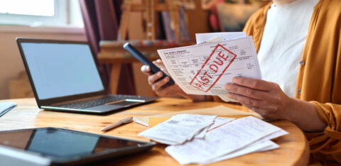 Woman reviewing an overdue bill while holding a smartphone at a table with a laptop and receipts.