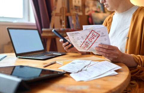 Woman reviewing an overdue bill while holding a smartphone at a table with a laptop and receipts.