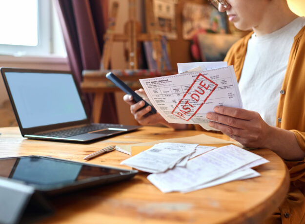 Woman reviewing an overdue bill while holding a smartphone at a table with a laptop and receipts.