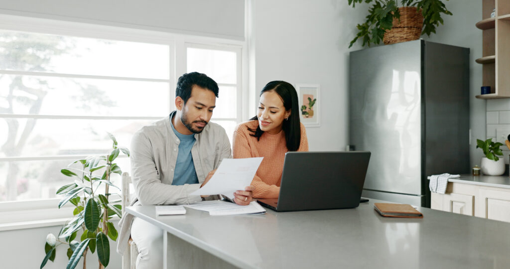 Couple reviewing financial documents together at a kitchen table with a laptop, discussing bills or household finances.