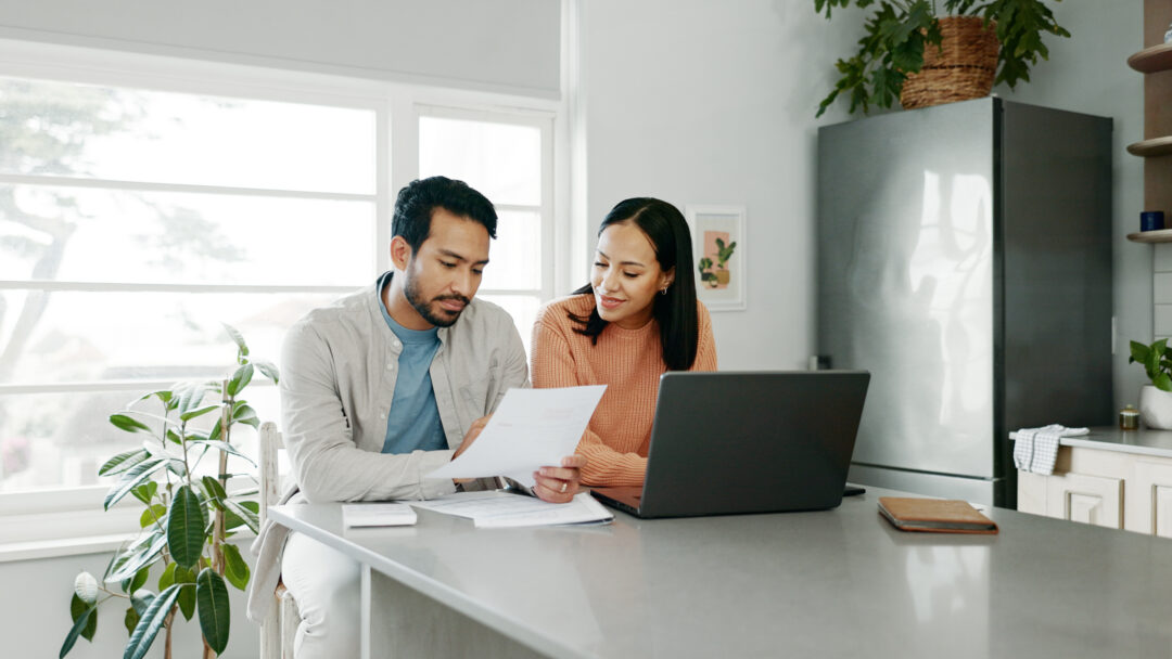 Couple reviewing financial documents together at a kitchen table with a laptop, discussing bills or household finances.