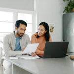 Couple reviewing financial documents together at a kitchen table with a laptop, discussing bills or household finances.