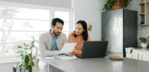 Couple reviewing financial documents together at a kitchen table with a laptop, discussing bills or household finances.