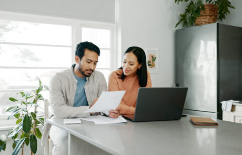 Couple reviewing financial documents together at a kitchen table with a laptop, discussing bills or household finances.
