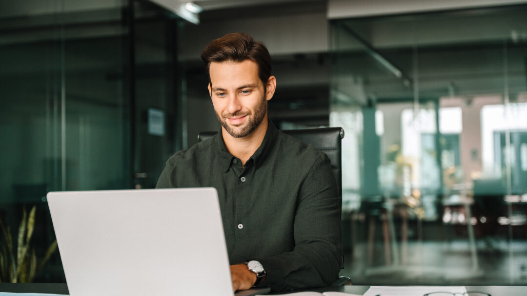 Happy latin hispanic young business man working on laptop computer in company office. Smiling European entrepreneur manager businessman using pc for communication, learning at workplace. Copy space