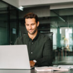 Happy latin hispanic young business man working on laptop computer in company office. Smiling European entrepreneur manager businessman using pc for communication, learning at workplace. Copy space