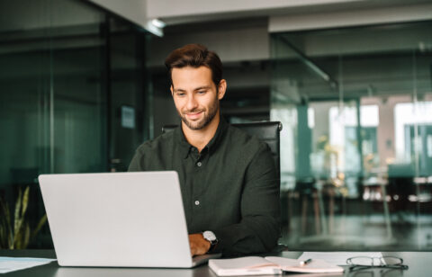 Happy latin hispanic young business man working on laptop computer in company office. Smiling European entrepreneur manager businessman using pc for communication, learning at workplace. Copy space