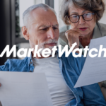 Older couple sitting on a couch reviewing paperwork together with a MarketWatch logo overlay.