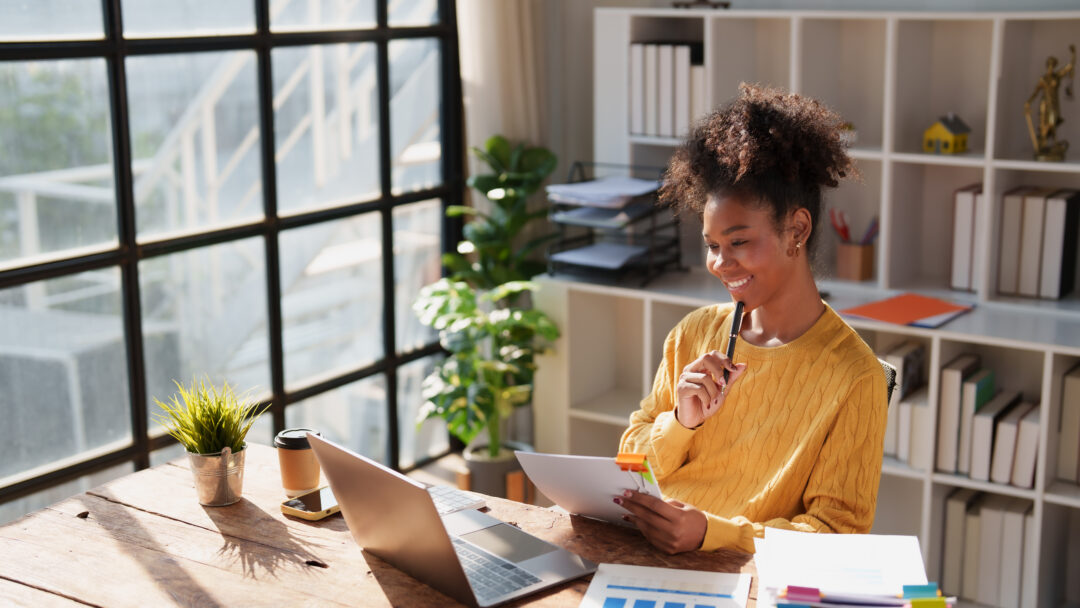 Young happy African American woman reviewing financial documents and planning strategy at her desk, with a laptop and organized files in a modern office environment