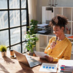 Young happy African American woman reviewing financial documents and planning strategy at her desk, with a laptop and organized files in a modern office environment