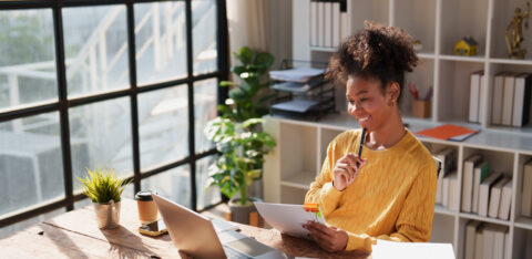 Young happy African American woman reviewing financial documents and planning strategy at her desk, with a laptop and organized files in a modern office environment