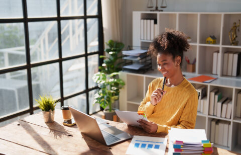 Young happy African American woman reviewing financial documents and planning strategy at her desk, with a laptop and organized files in a modern office environment