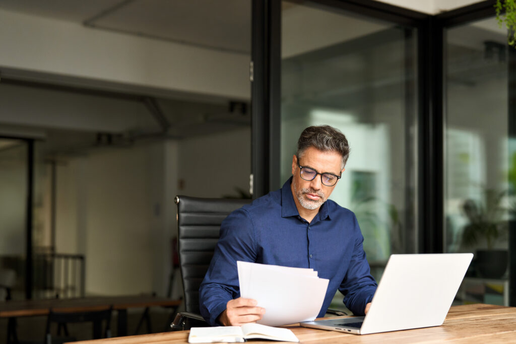 Busy mature professional business man using laptop reading document at work.