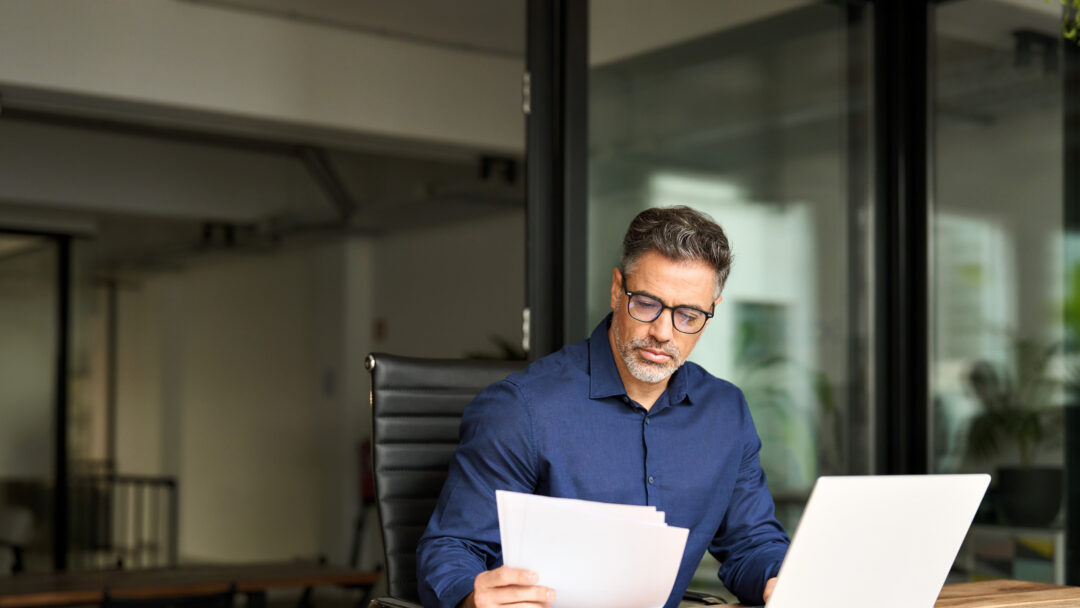 Busy mature professional business man using laptop reading document at work.