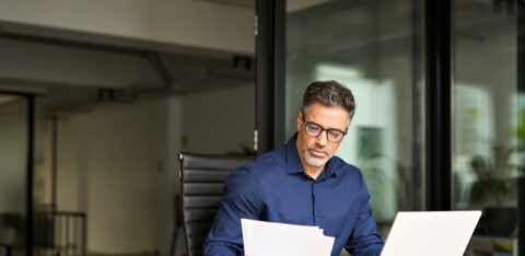 Busy mature professional business man using laptop reading document at work.