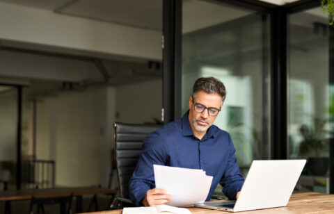 Busy mature professional business man using laptop reading document at work.